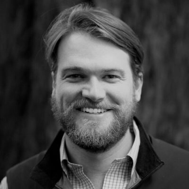 The image shows a black and white portrait of a man with a beard and short hair. He is smiling and looking at the camera. He is wearing a collared shirt and a vest. The background is blurred, suggesting an outdoor setting.